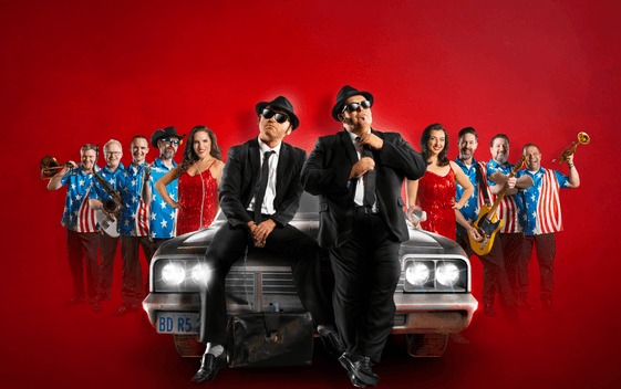 The cast of Chicago Blues Brothers are arranged either side of a car, with the two lead chracters sat on the bonnet of the car, in front of a red background