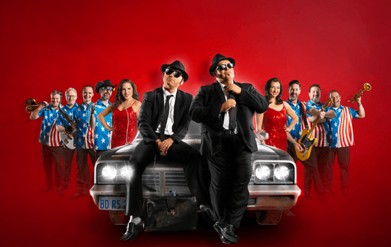 The cast of Chicago Blues Brothers are arranged either side of a car, with the two lead chracters sat on the bonnet of the car, in front of a red background