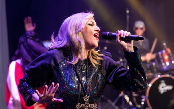 A performer impersonating Madonna is wearing a blakck outfit and singing into a microphone on stage