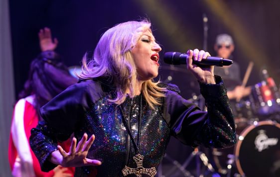 A performer impersonating Madonna is wearing a blakck outfit and singing into a microphone on stage