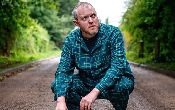 Comedian Miles Jupp is crouched down on a muddy path with bare feet, wearing blue checked pyjamas with a concerned expression on his face
