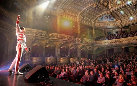 A performer, impersonating Freddie Mercury, stands at the front of the stage in a white and red suit with one fist in the air, facing an audience in an old fashioned theatre hall.