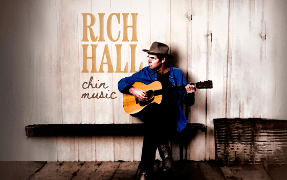 Rich Hall sits with a guitar, wearing a cowboy hat against a wooden backdrop. The words "Rich Hall Chin Music" appear next to him