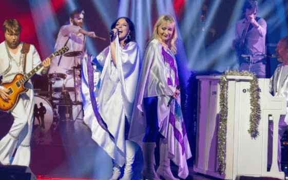 The lead performers and band of Abbamania are shown in 70's costumes signing and playing instruments surrounded by colourful light beams