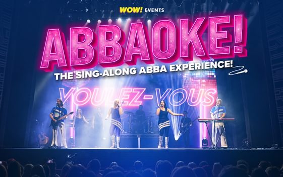 The show title "Abbaoke" is shown in bright pink above an image of 4 performers, dressed like ABBA, performing on a stage. The words "Voulez Vous" are shown behind them in neon lights.