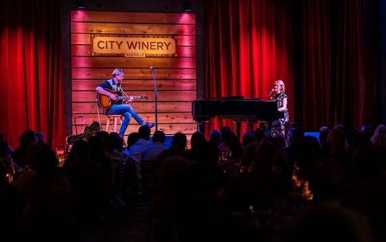 Two performers on a stage, performing as Carole King sat at a baby grand piano and James Taylor playing guitar.