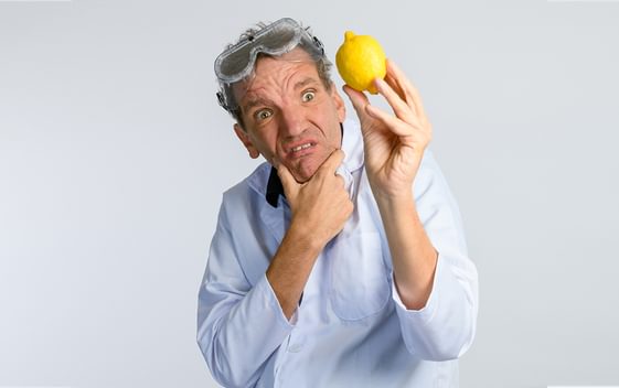 Henning Wehn with a confused facial expression, wearing clear goggles on his head and holding a lemon up in front of him