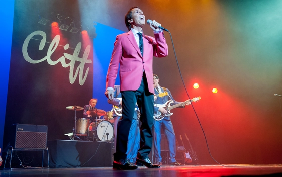 Jimmy Jemain performing as Cliff Richard, wearing a red jacket. There is a live band in the background and a large sign which says "Cliff" at the back of the stage.