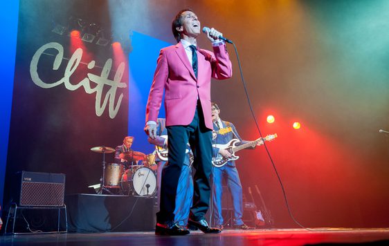 Jimmy Jemain performing as Cliff Richard, wearing a red jacket. There is a live band in the background and a large sign which says "Cliff" at the back of the stage.