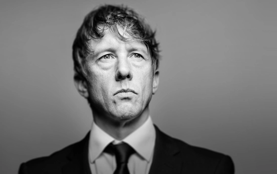 A black and white image of Jonathan Pie wearing a suit and tie