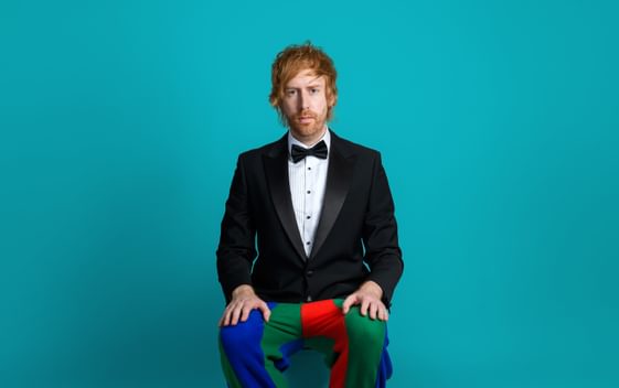 Mark Simmons is sat in front of a blue background wearing a black tuxedo, white shirt and bowtie, with a jesters trousers and shoes.