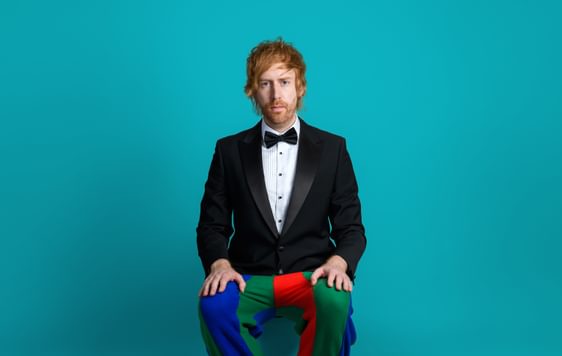 Mark Simmons is sat in front of a blue background wearing a black tuxedo, white shirt and bowtie, with a jesters trousers and shoes.