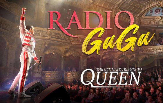 A performer, impersonating Freddie Mercury, stands at the front of the stage in a white and red suit with one fist in the air, facing an audience in an old fashioned theatre hall.