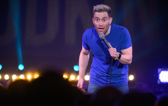 Simon Brodkin is shown on stage wearing a blue t-shirt and holding a microphone.
