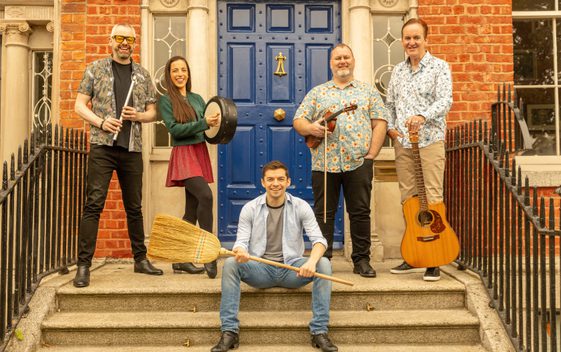 The Irish House Party band are shown sat on concrete steps with a blue door behind them. They are holding instruments and one person is holding a broom.