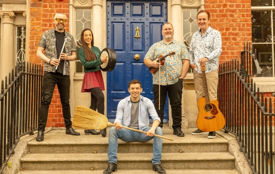 The Irish House Party band are shown sat on concrete steps with a blue door behind them. They are holding instruments and one person is holding a broom.