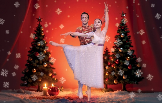 A male and a female ballet dancer are dancing on stage with a backdrop of snow, Christmas trees and candlelight.