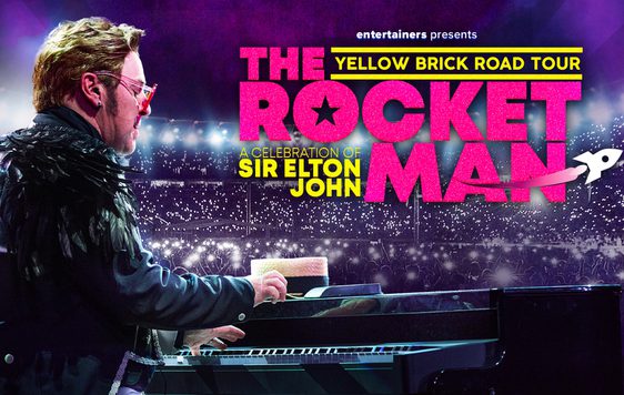 A performer dressed as Elton John with black feathers on his shouulders, is sitting at a piano with a large crowd in the background holding up lights.