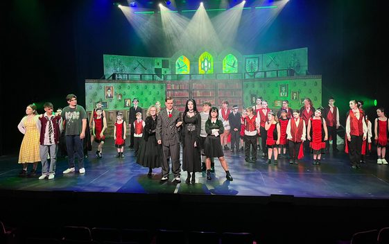 A cast of young performers on stage as the Addams Family with school children stood behind. The set is lit with green light behind them.