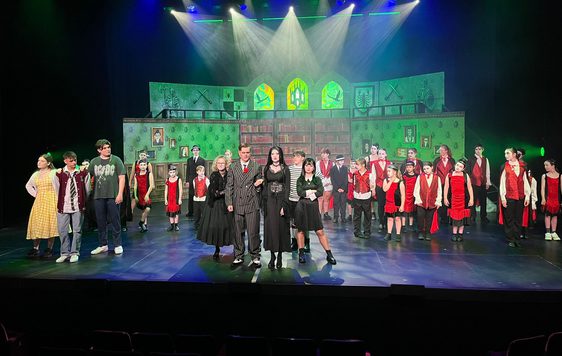 A cast of young performers on stage as the Addams Family with school children stood behind. The set is lit with green light behind them.