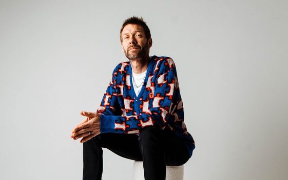 Tom Meighan of "Kasabian" is sitting on a white stool with his hands pressed together, wearing black jeans a white t shirt and blue patterned cardigan.