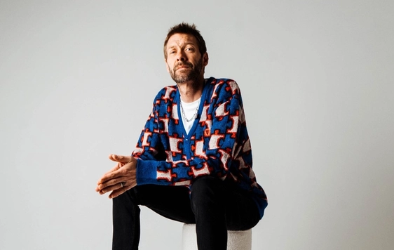 Tom Meighan of "Kasabian" is sitting on a white stool with his hands pressed together, wearing black jeans a white t shirt and blue patterned cardigan.