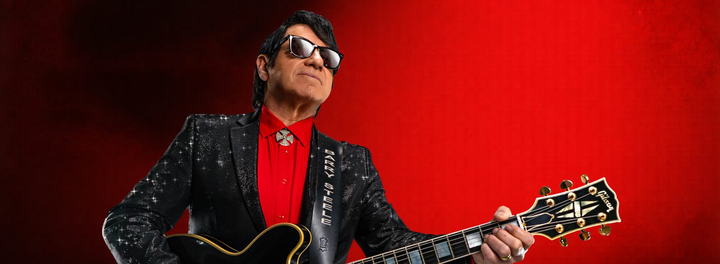 Barry Steele posing as Roy Orbison with a guitar against a red background