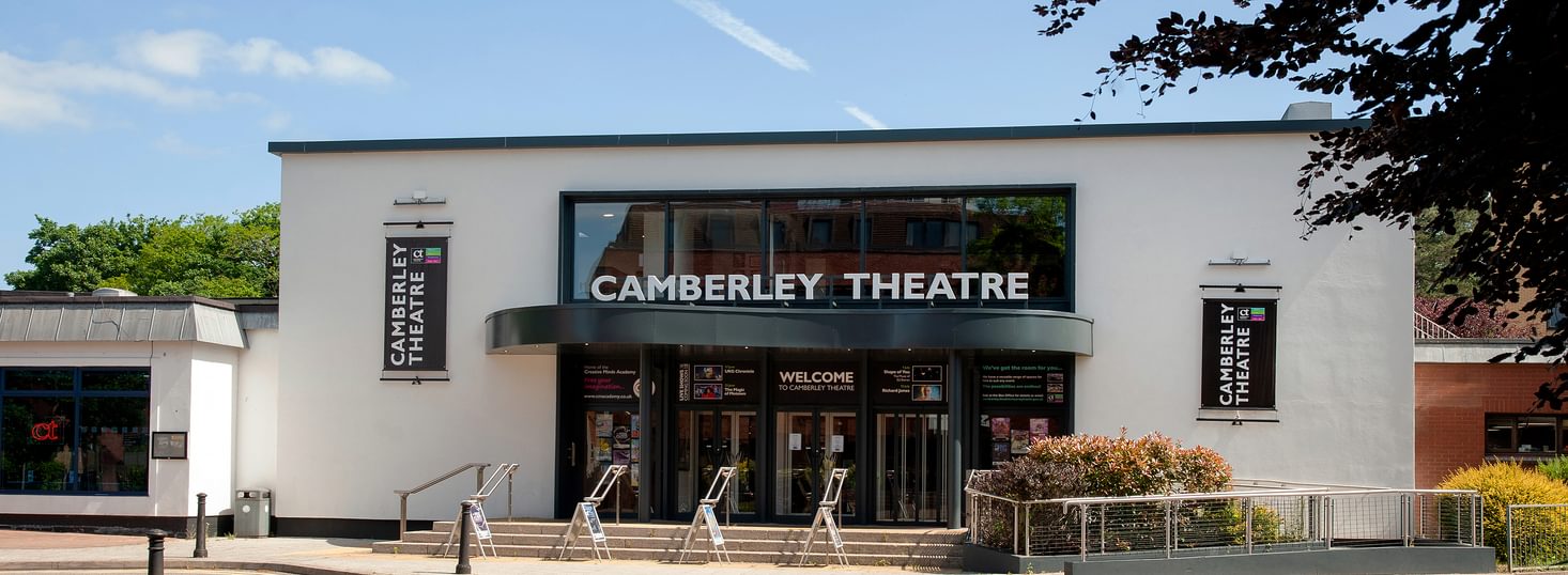 A view of the Camberley Theatre frontage - a building with white walls and a dark grey canopy above the front doors with a sign reading "Camberley Theatre".