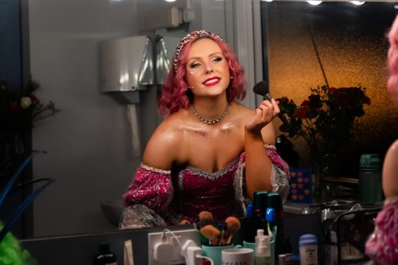 A performer, dressed as a fairy godmother with pink hair is applying make-up in a dressing room mirror
