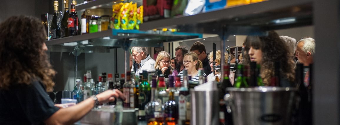 The back of the bar at Camberley Theatre with bottles of spirits, an ice bucket and a mirror along the back. A member of staff is preparing a drink and customers can be seen in the mirror's reflection.