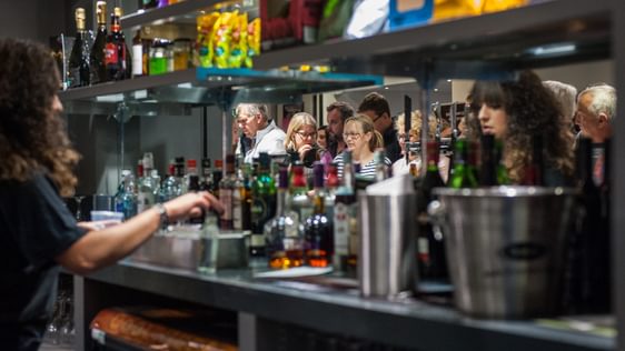 The back of the bar at Camberley Theatre with bottles of spirits, an ice bucket and a mirror along the back.  A member of staff is preparing a drink and customers can be seen in the mirror's reflection.
