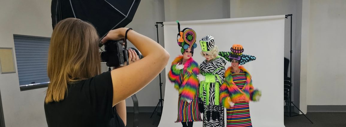 The ugly stepsisters and baroness on a white backdrop having a photoshoot. In the foreground the photographer is taking a picture of them.