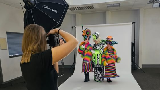 The ugly stepsisters and baroness on a white backdrop having a photoshoot. In the foreground the photographer is taking a picture of them.