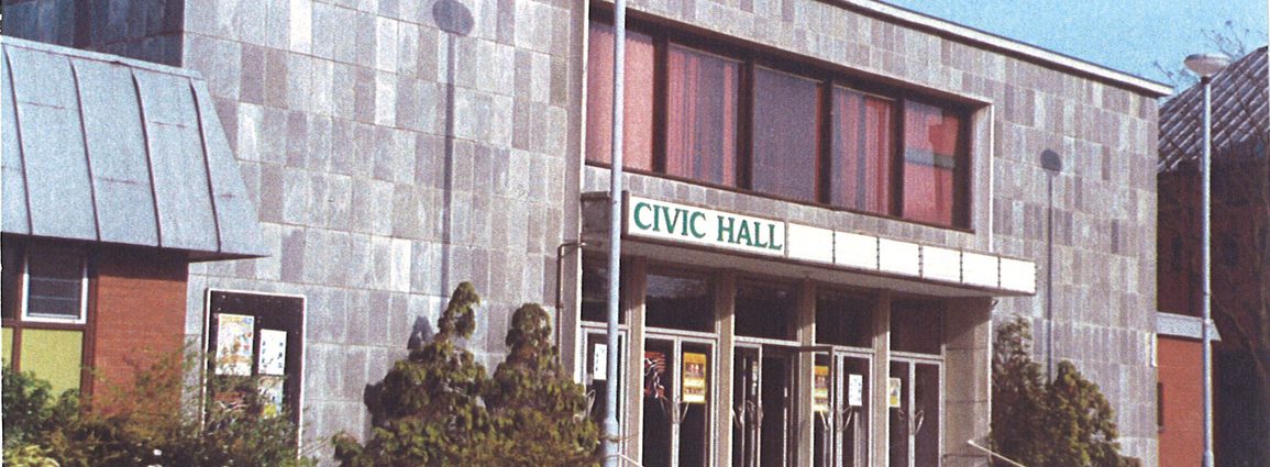A picture of the Camberley Civic Hall, clad with large grey tiles, with foliage along the front and steps up to the front of the building
