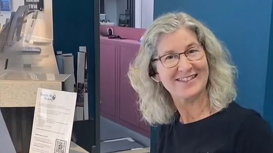 A box office assistant with a welcoming smile. She is wearing a black Camberley Theatre uniform and sitting behind the box office