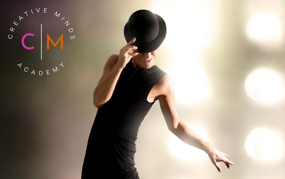 A performer in a sleeveless black top, wearing a black bowl hat tipped forward and they are dancing wth stage lights behind